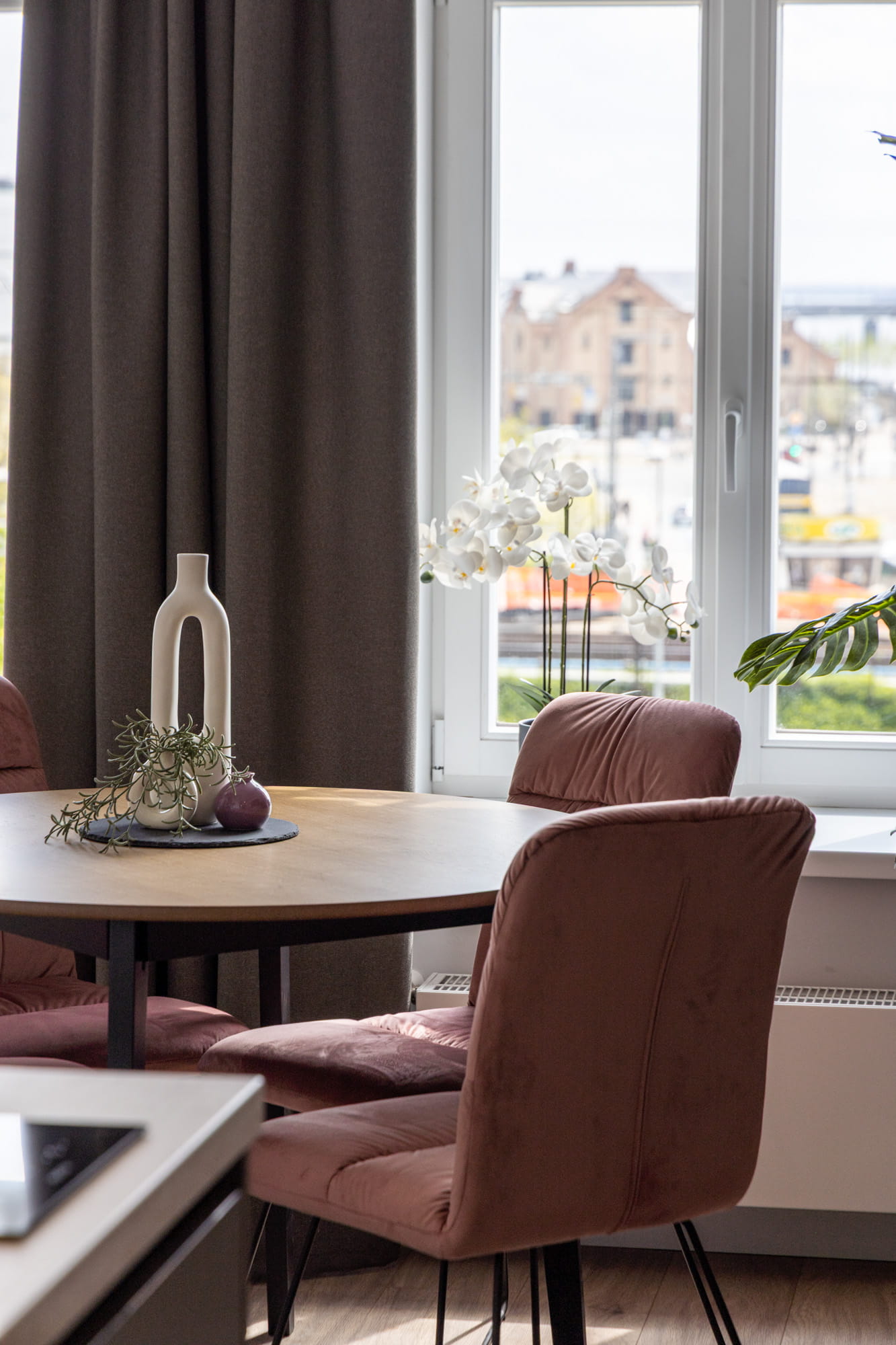 Apaļa koka ēdamgalds ar trīs rozā samta krēsliem pie loga ar baltām ziedu dekorācijām un tumšu aizkaru fonā.