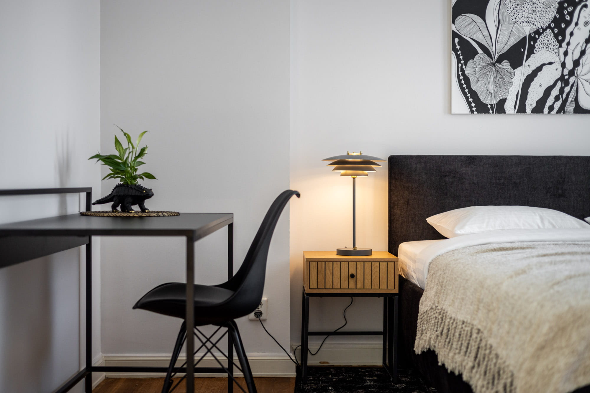 Mūsdienīga guļamistaba ar melnu biroja krēslu, melnu galda virsmu ar dinosaurusa formas puķupodu, naktsgaldiņu ar lampu un melnu gultas galvgali.