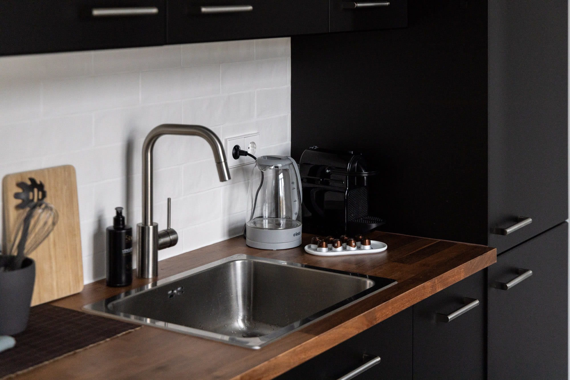 Mūsdienīga virtuves izlietne ar metāla jaucējkrānu, elektrisko tējkannu, kafijas automātu un kapsulām uz koka virsmas.