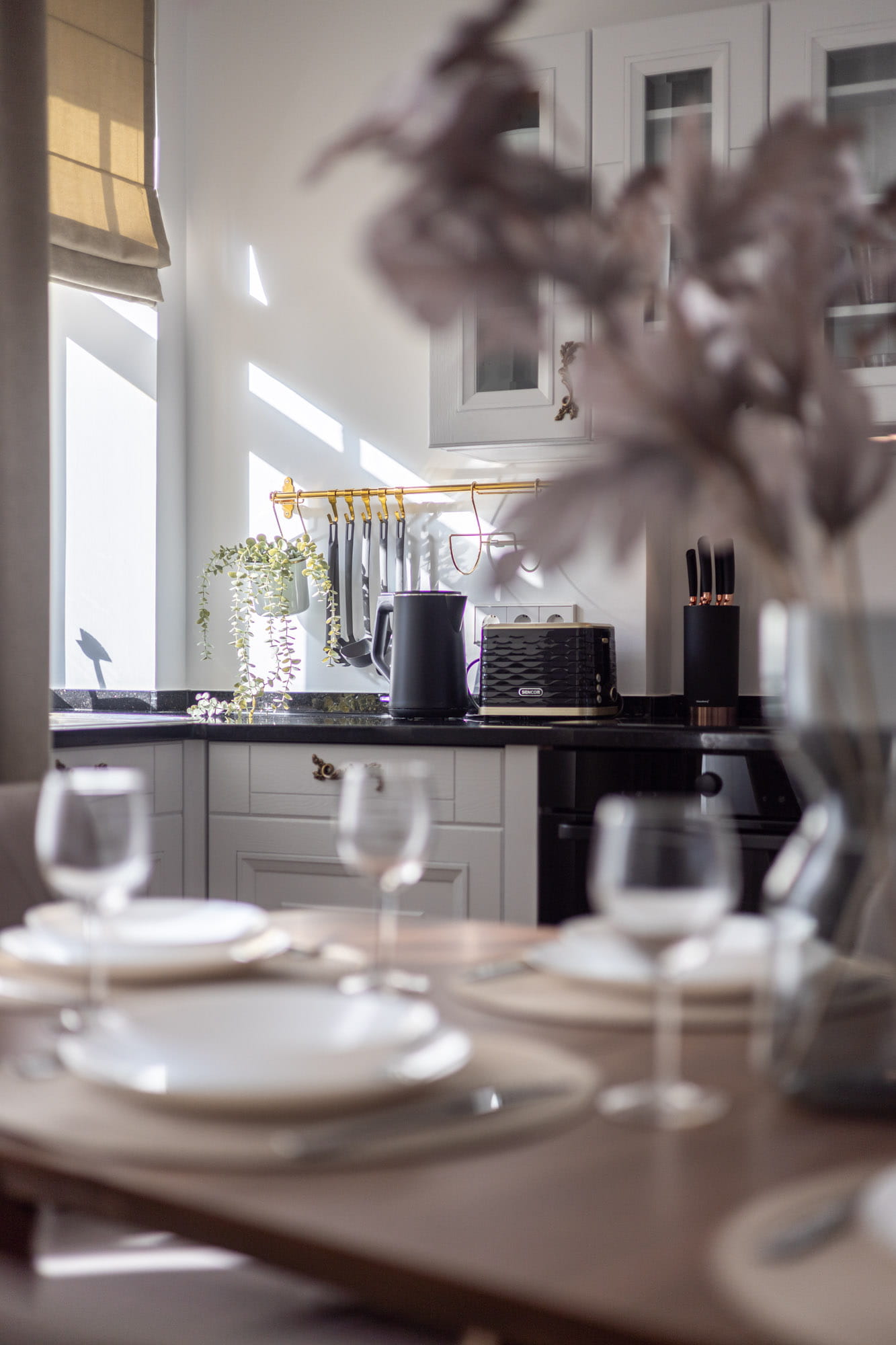 Modernā virtuve ar baltām skapīšu durvīm, melnu galda virsmu, elektrisko tējkannu, maiznīcu, nažu komplektu un galdā klātiem traukiem ar glāzēm.