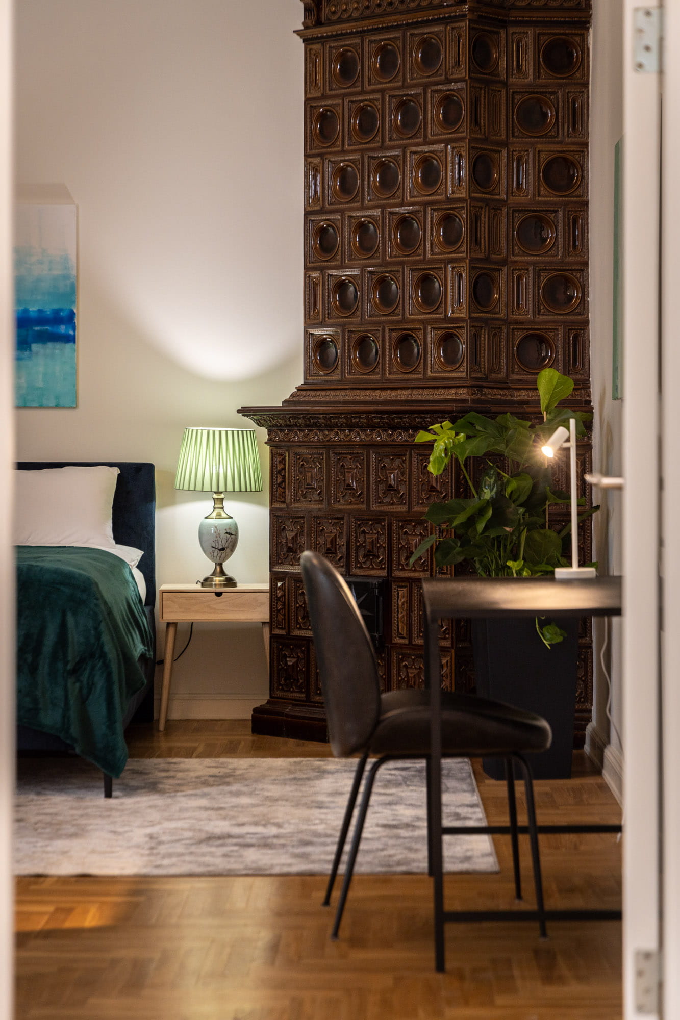 Darba zona ar melnu krēslu un galdu, pie sienas ir liela brūna vēsturiska keramikas krāsns, blakus zaļa istabas augs un naktsgaldiņš ar lampu pie gultas ar zaļu segu.