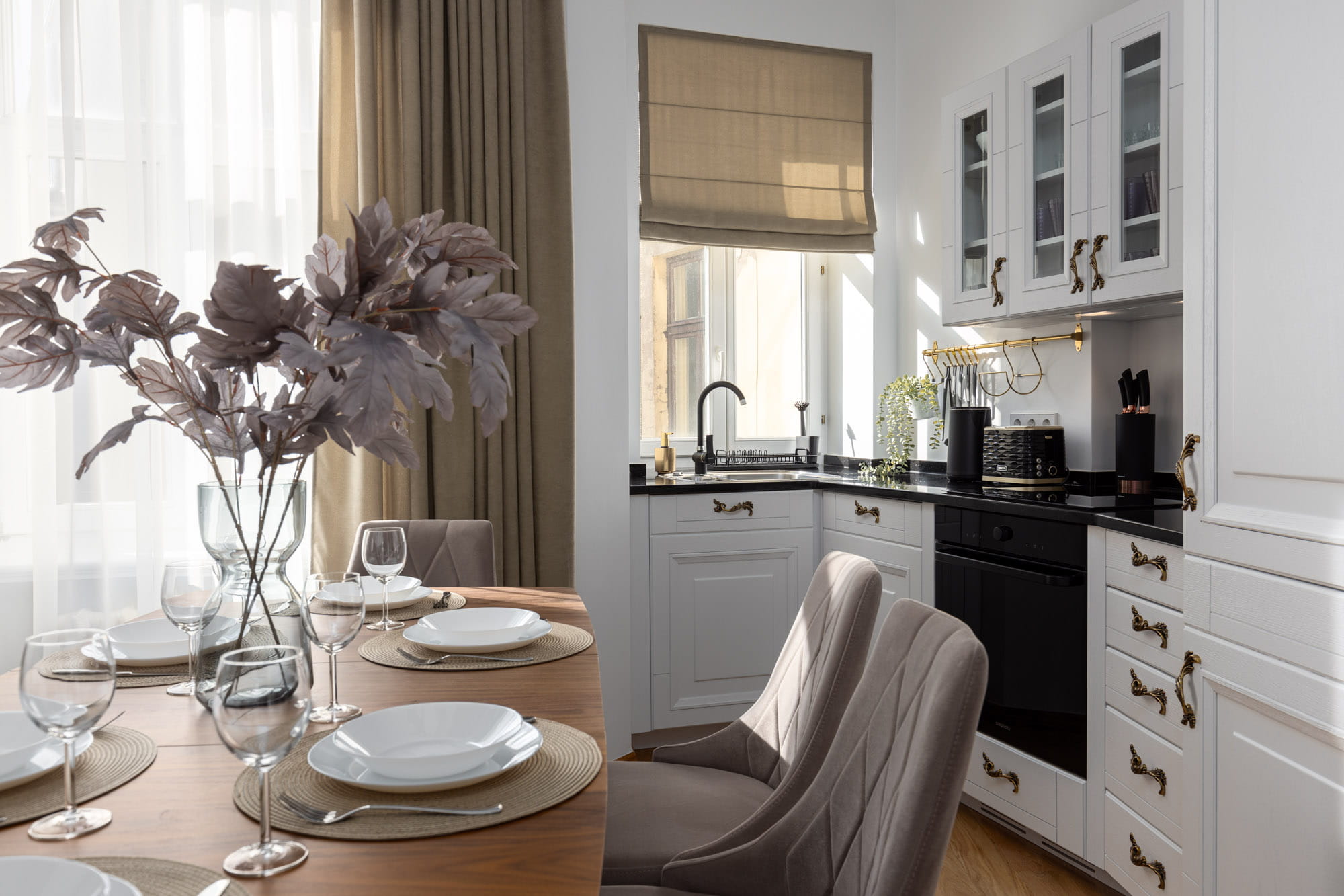 Mūsdienīga virtuve ar baltām skapīšiem, melnu izlietni, pelēkām atzveltnes krēsliem un koka ēdamgaldu ar baltām šķīvēm un glāzēm.