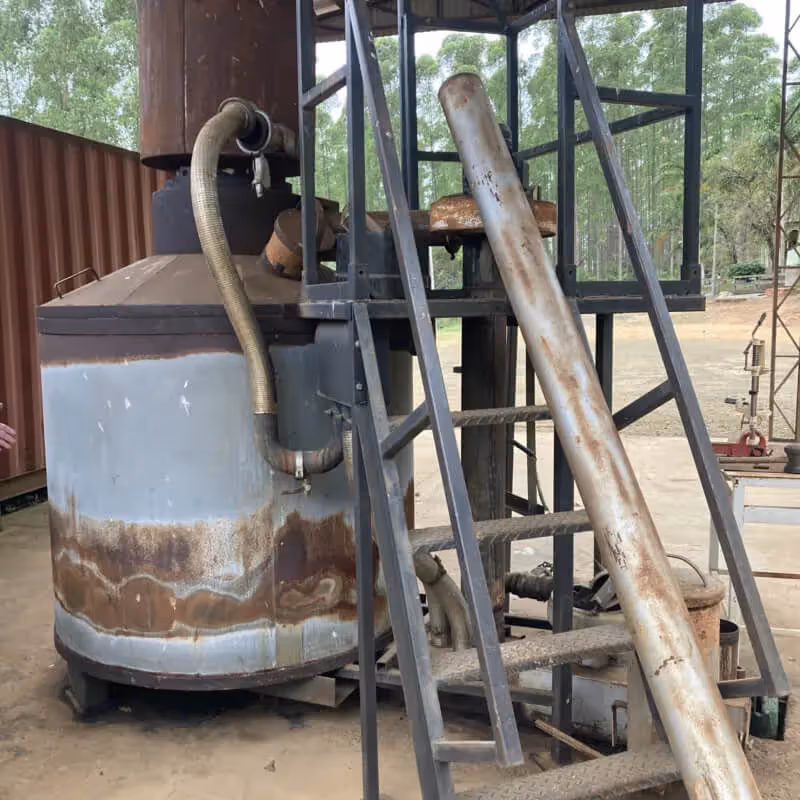 Industrial metal machine with pipes and a staircase, set outdoors near trees and a shipping container.