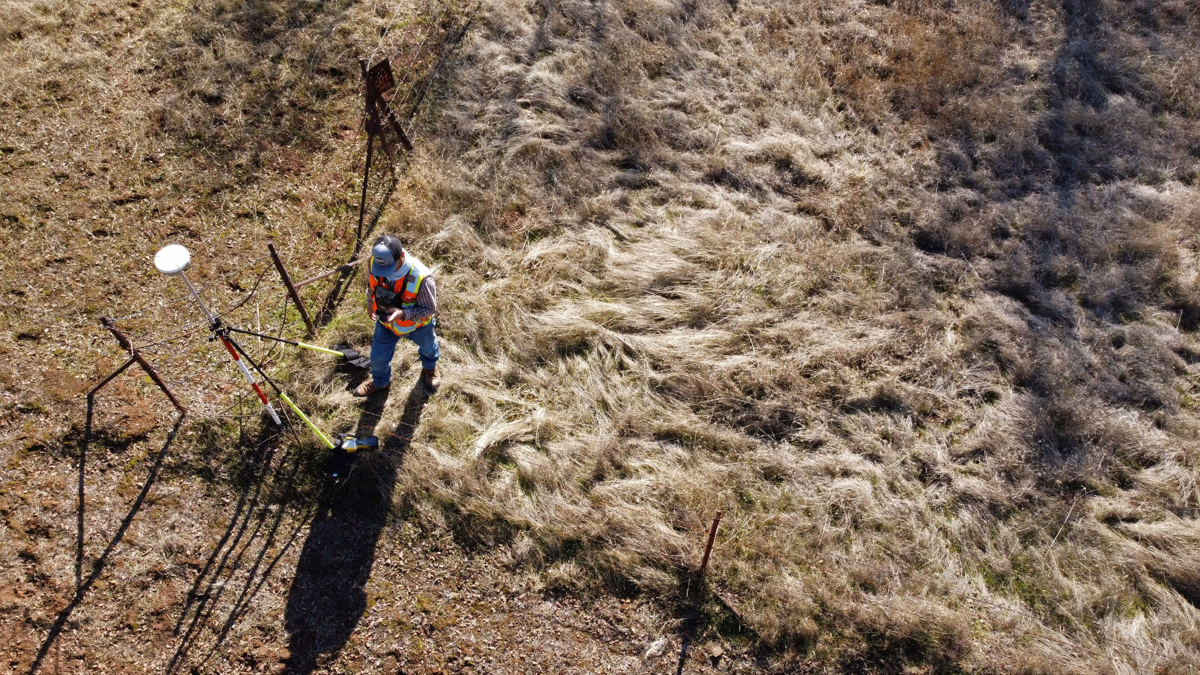 Surveyor wearing a reflective vest and cap standing on grassy terrain using surveying equipment with a tripod.