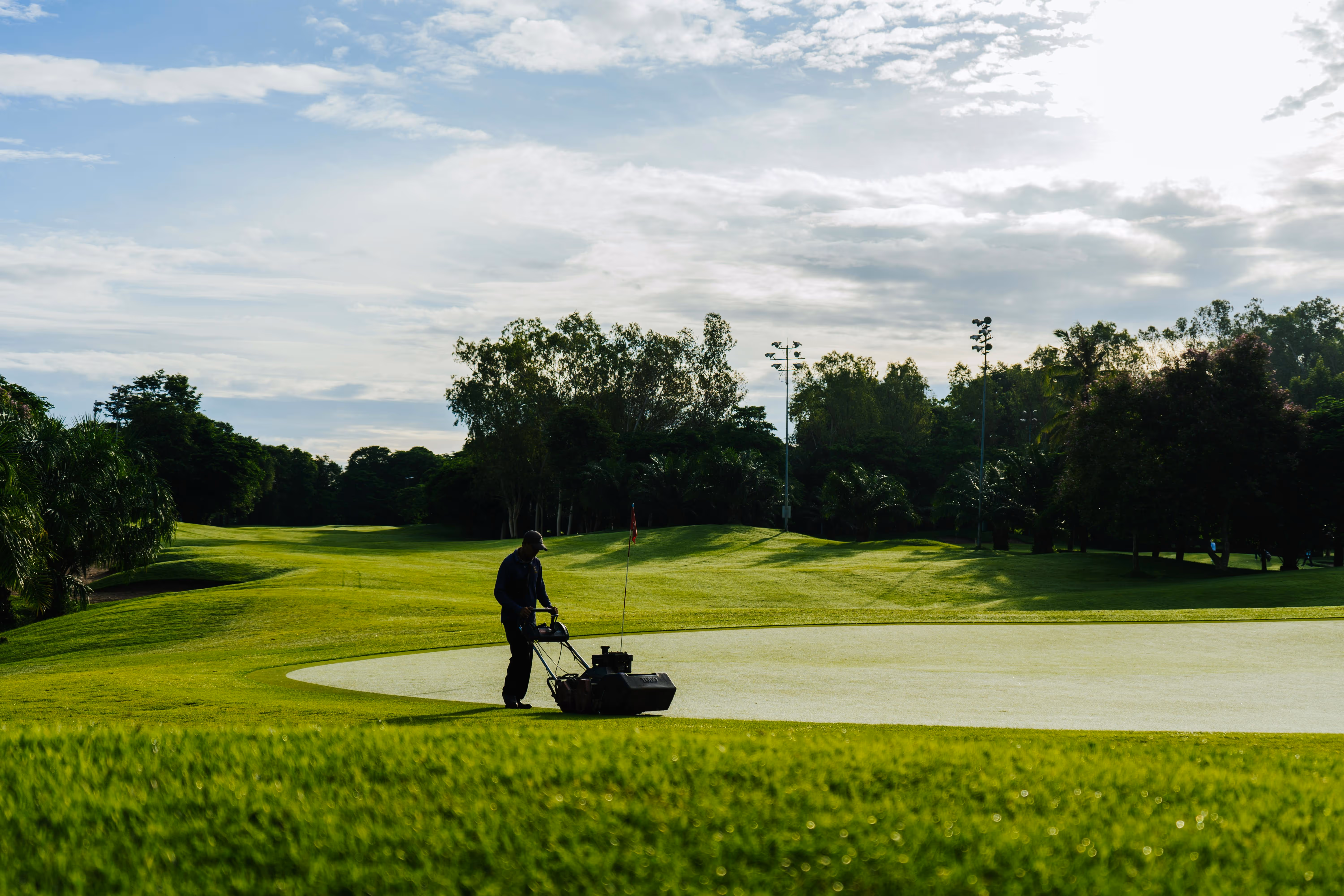 Person mowing grass near a putting green on a golf course under a cloudy sky.