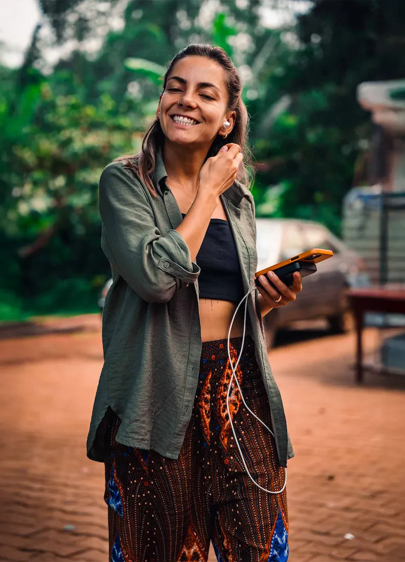 Smiling woman outdoors wearing patterned pants and a green shirt, holding a phone and adjusting an earbud.
