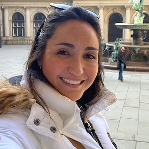 Smiling woman with sunglasses on her head wearing a white jacket, standing in an outdoor courtyard with a fountain in the background.