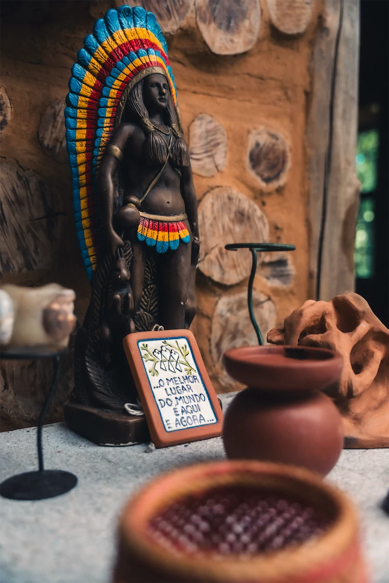 Statue of an indigenous woman with a colorful feathered headdress standing next to a sign reading 'O melhor lugar do mundo é aqui e agora' in a rustic setting.