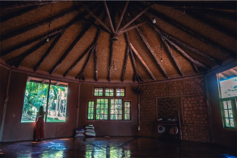 Interior of a rustic round room with a wooden ceiling and large windows showing greenery outside, with a person standing by one window.