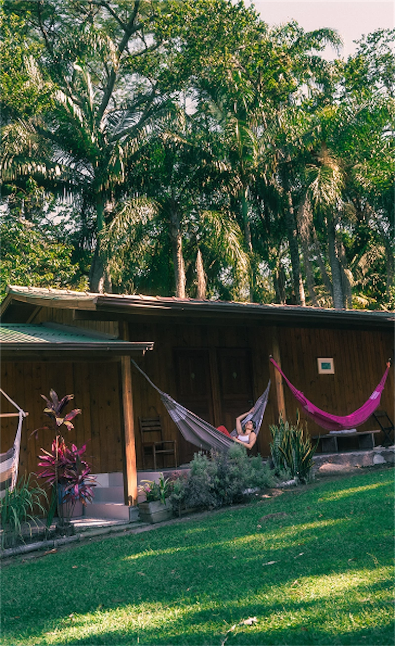 Woman relaxing in a hammock on the porch of a wooden cabin surrounded by palm trees and green lawn.