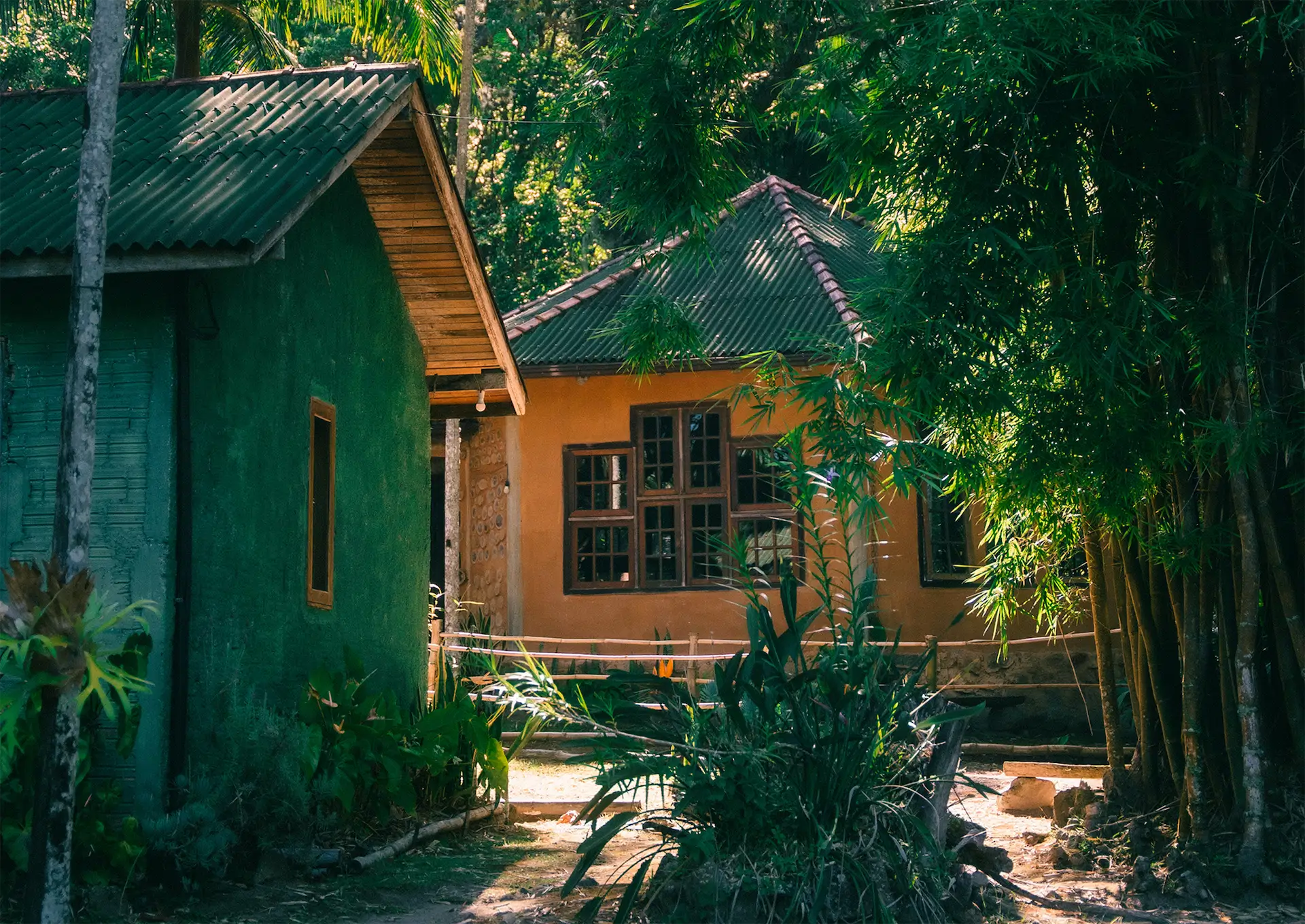 Two small houses surrounded by lush greenery and bamboo plants with sunlight filtering through the trees.