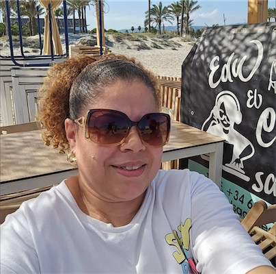 Woman wearing large sunglasses and a white shirt, smiling while sitting outdoors near a beach with palm trees and a blackboard with white text.