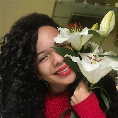 Woman with long curly hair and red lipstick smiling while holding white lilies close to her face.