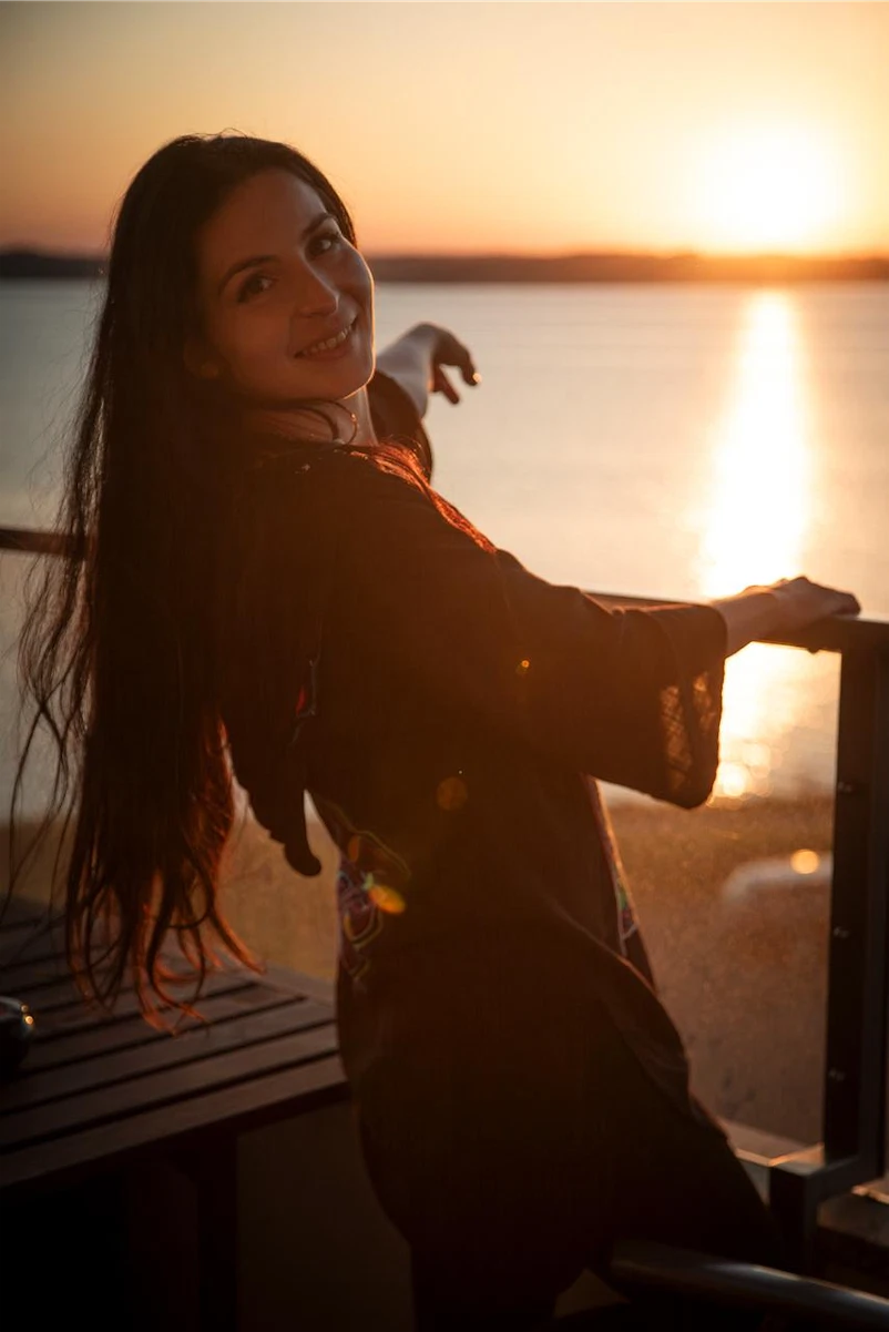 Smiling woman outdoors wearing patterned pants and a green shirt, holding a phone and adjusting an earbud.