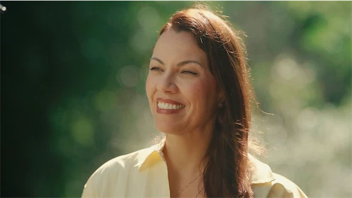 Smiling woman with sunglasses on her head wearing a white jacket, standing in an outdoor courtyard with a fountain in the background.
