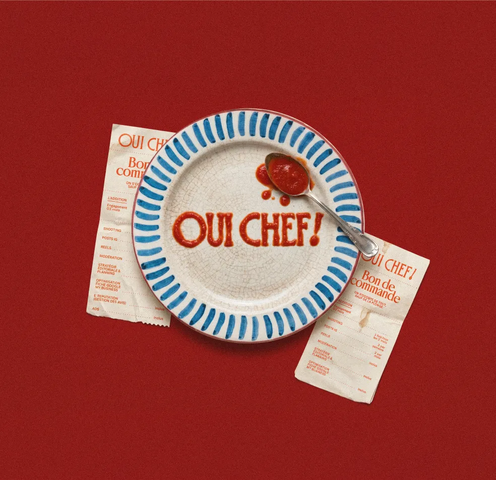 Assiette avec les mots 'OUI CHEF!' écrits en ketchup et une cuillère dessus, fond rouge.