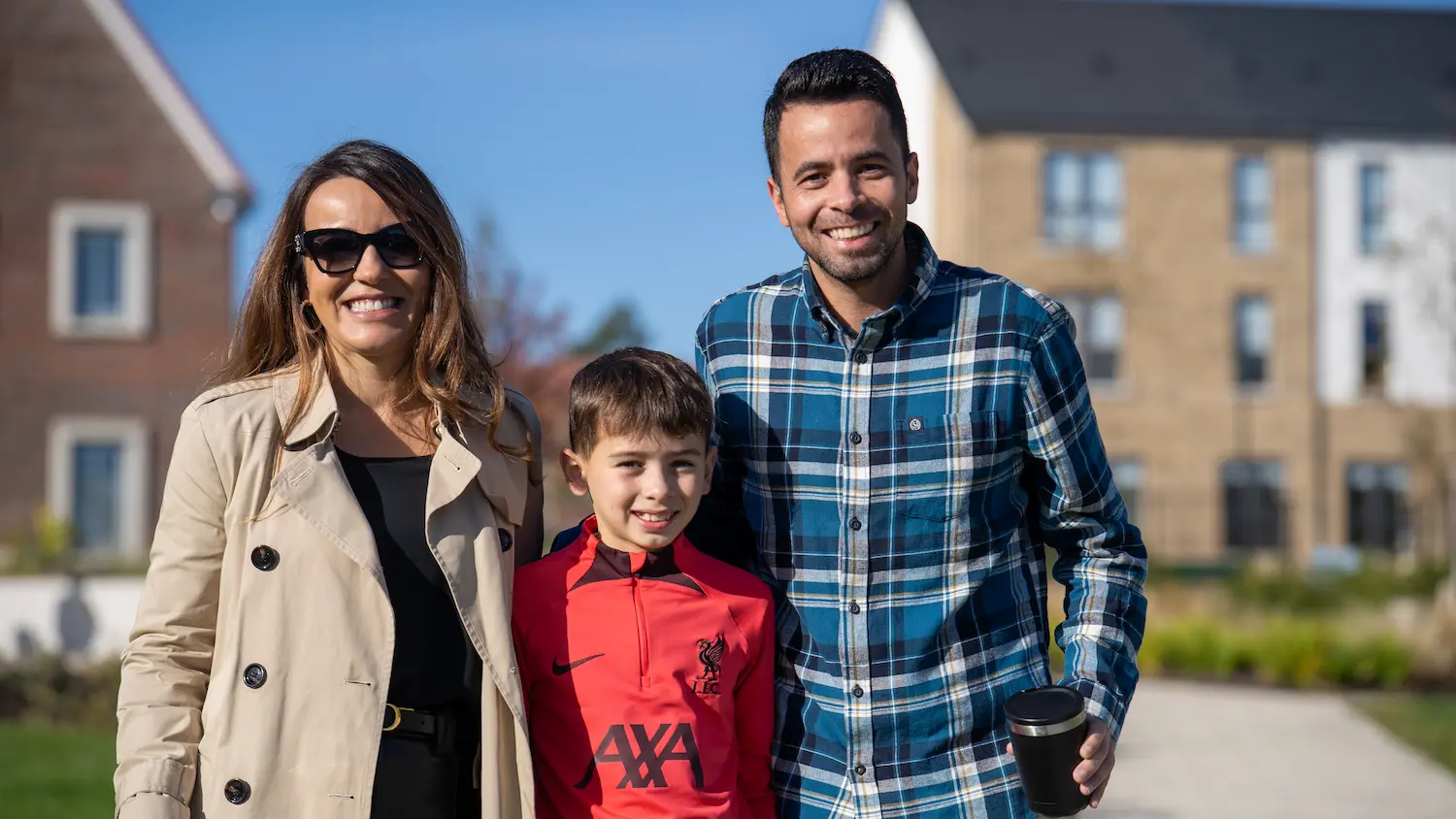 Smiling family outdoors in sunny neighborhood with woman in beige coat and sunglasses, boy in red jacket, and man in blue plaid shirt holding a travel mug.