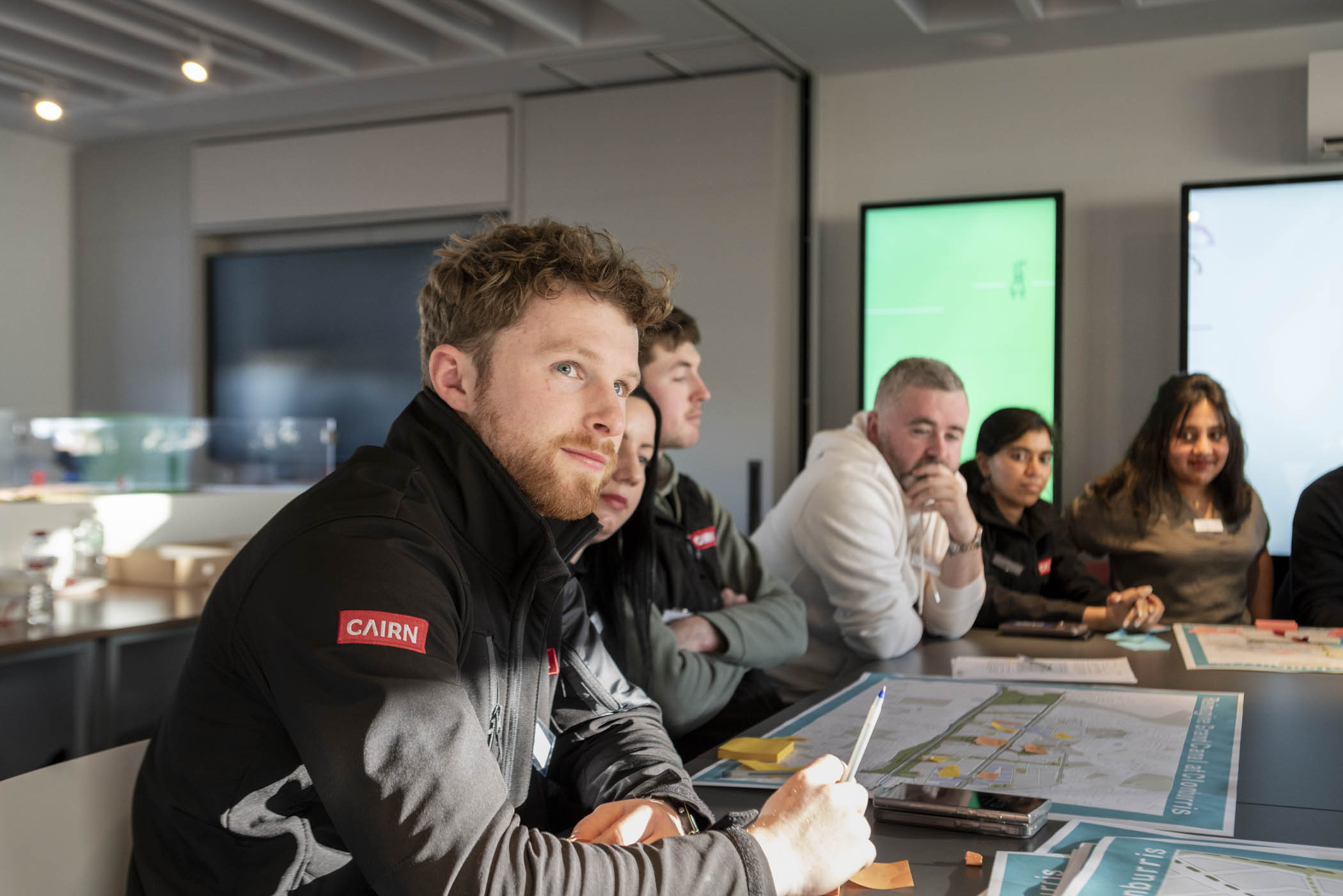 A group of six diverse people sitting around a table with maps and notes, engaged in discussion in a modern meeting room.