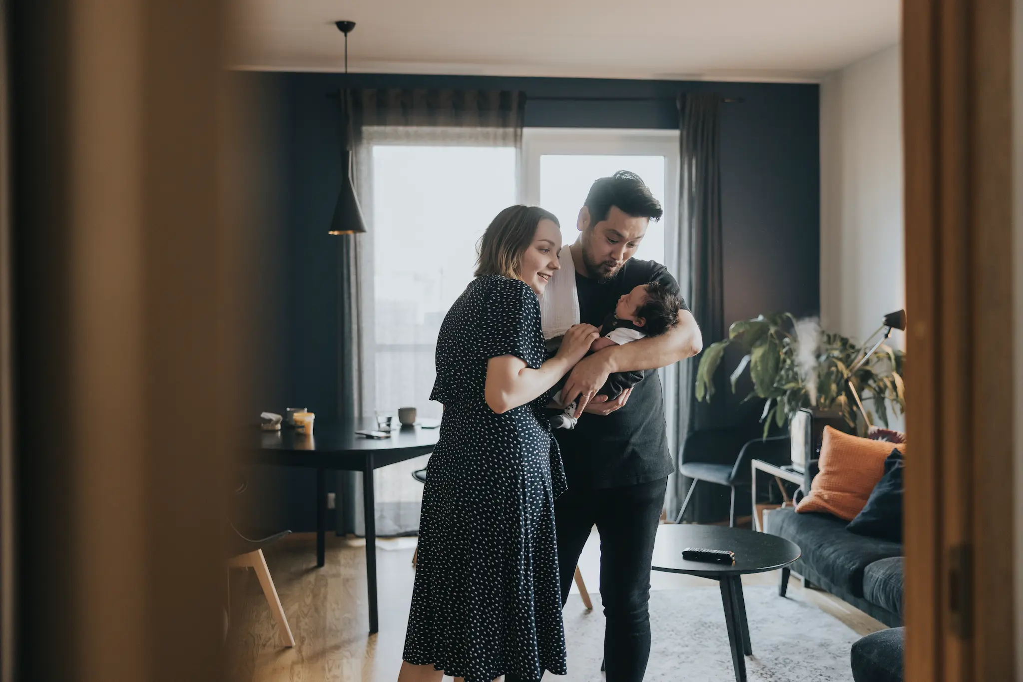 Couple standing in a cozy living room, smiling and looking at their baby who is being held by the man.