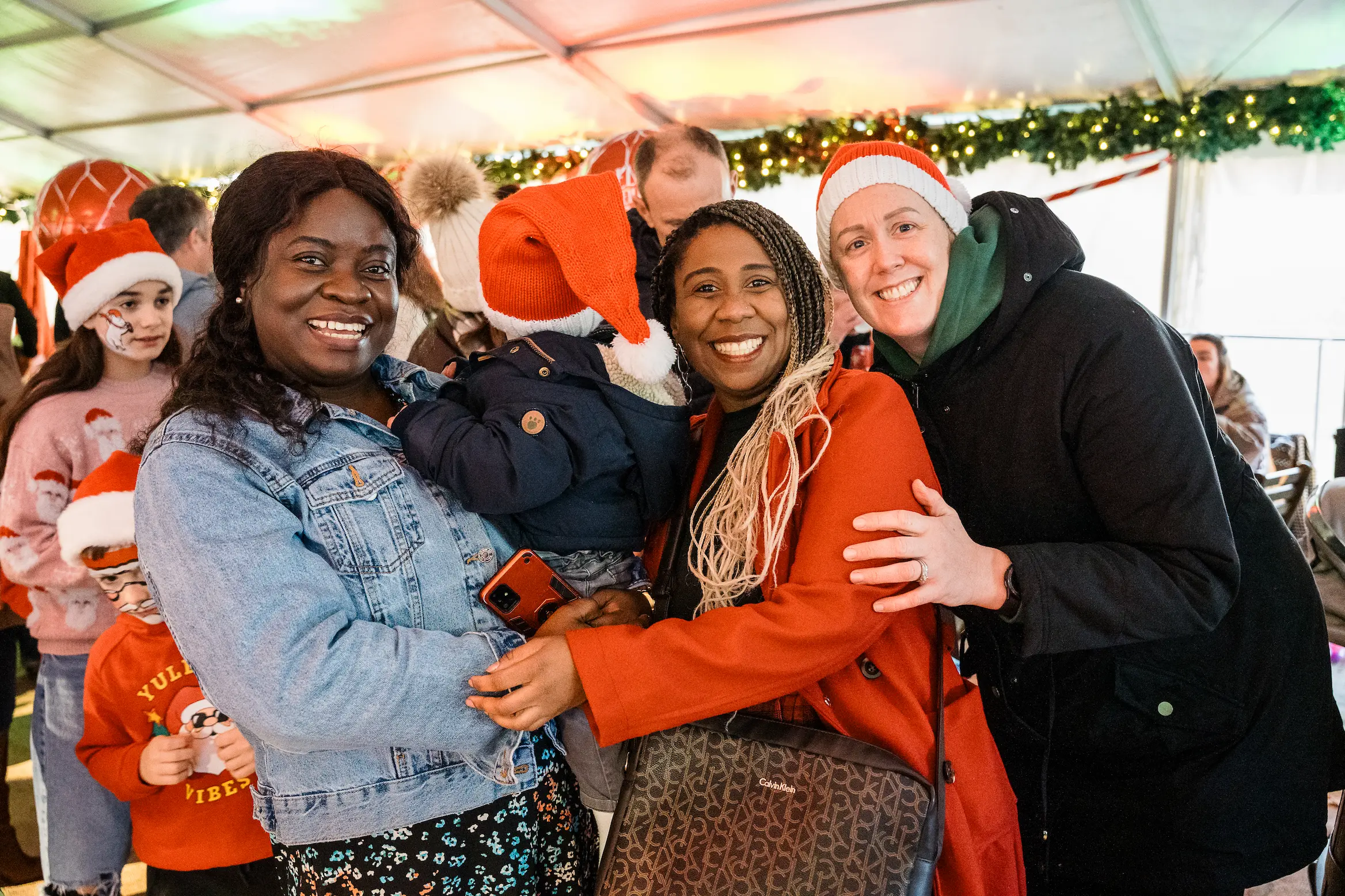 Three smiling adults and a child wearing Santa hats at a festive indoor event with holiday decorations.