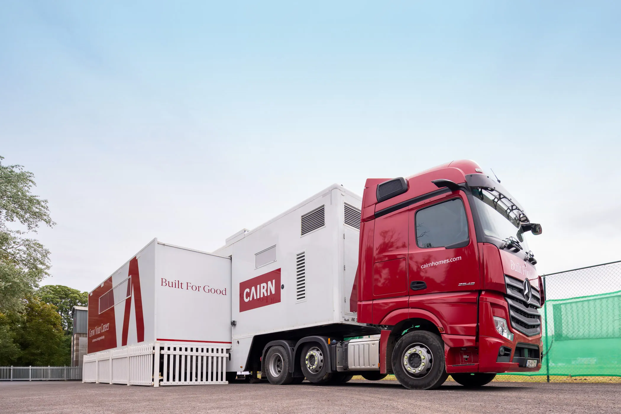 Red and white Cairn-branded truck trailer with the phrases 'Built For Good' and 'Grow Your Career' parked on a paved area near a fence.