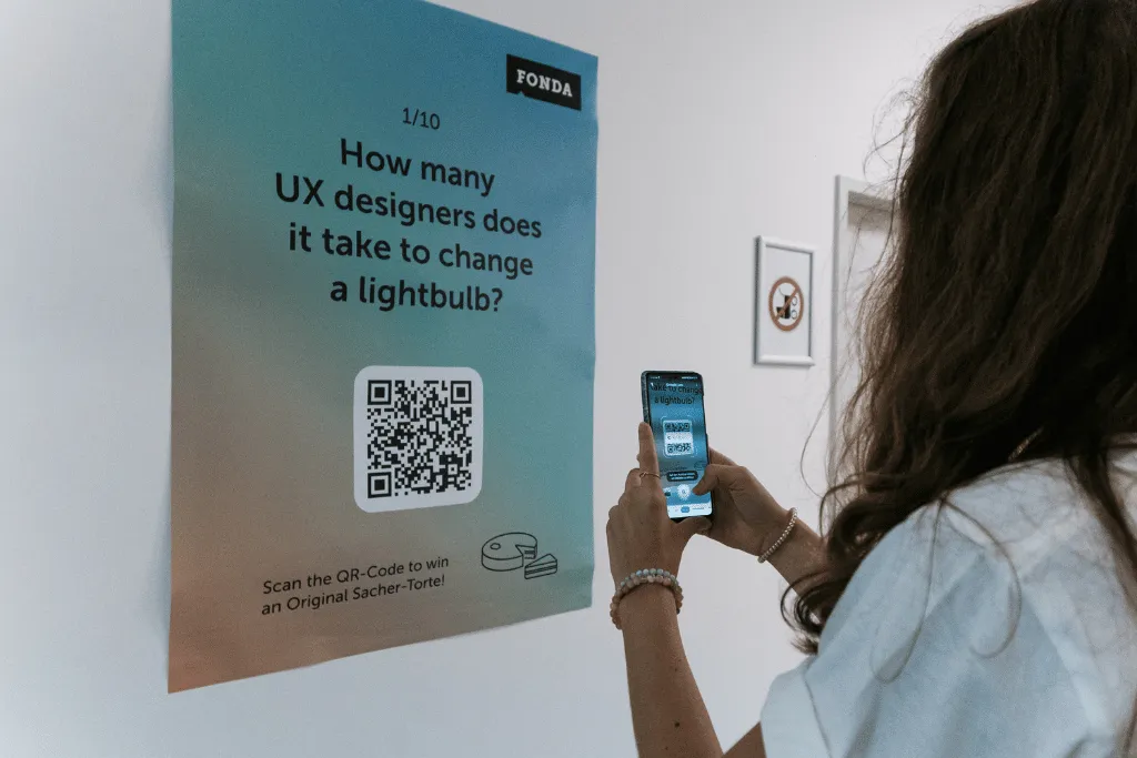 A woman scanning a QR code on a poster that says "How many UXers does it take to change a lightbulb?".