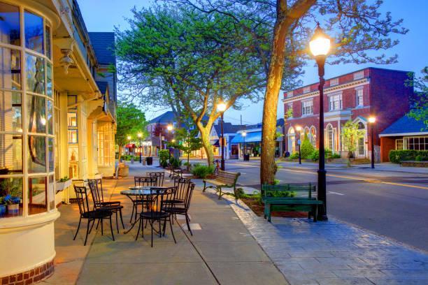 An image of downtown Falmouth showing 2 bistro tables and 8 chairs on the sidewalk lined with trees and lamp posts and businesses in the background.