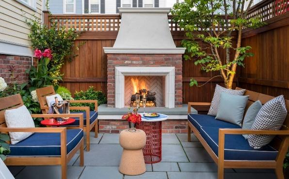 An image of an outdoor sofa facing 2 chairs and side table with a lit fireplace and wooden privacy wall in the background.