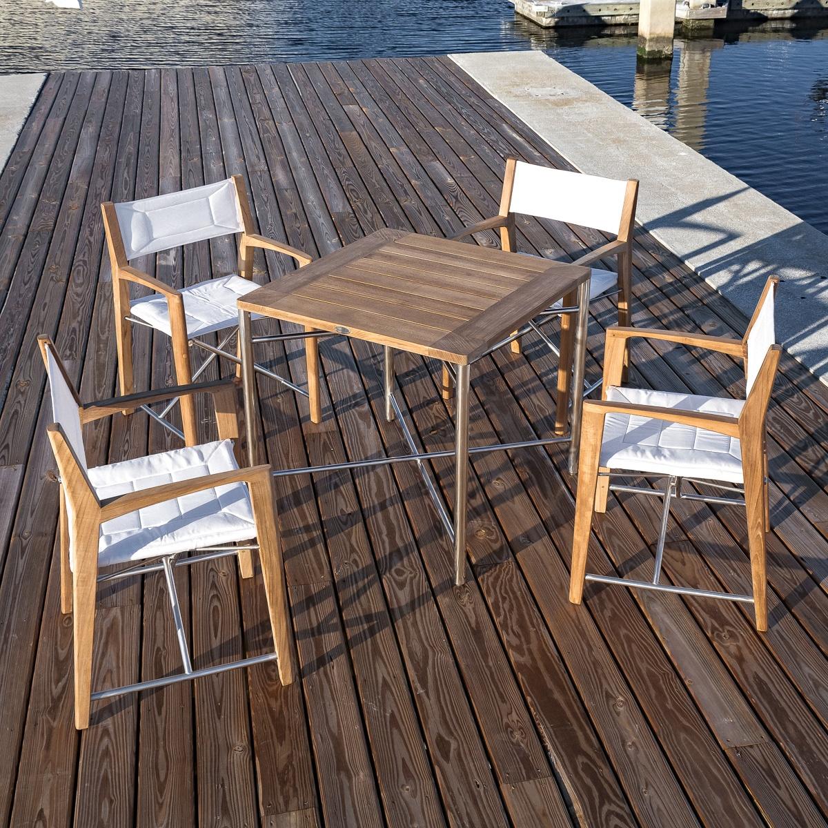 An image of 70459 Odyssey 5 Piece folding teak and stainless steel dining set of Odyssey 32 inch square dining table and 4 Odyssey folding chairs on a boat dock next to a boat marina.