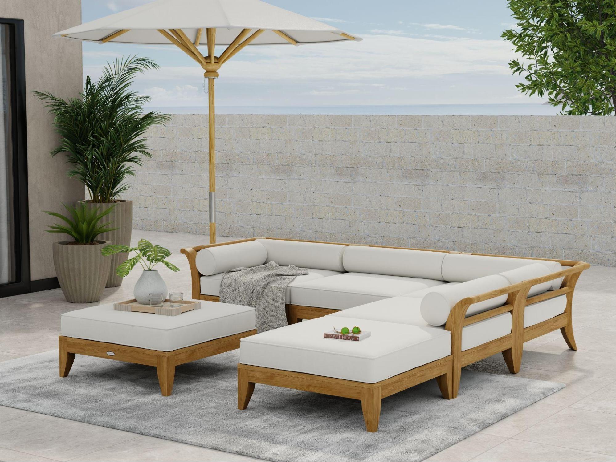 An image of 70121 Aman Dais 6 piece teak sectional set next to an umbrella on a stone patio with vase and 2 glasses on a tray on top of ottoman and book with sunglasses on cushion plant against wall