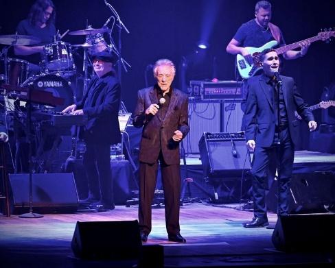 Frankie Valli on a stage with a microphone and a group of men playing instruments
