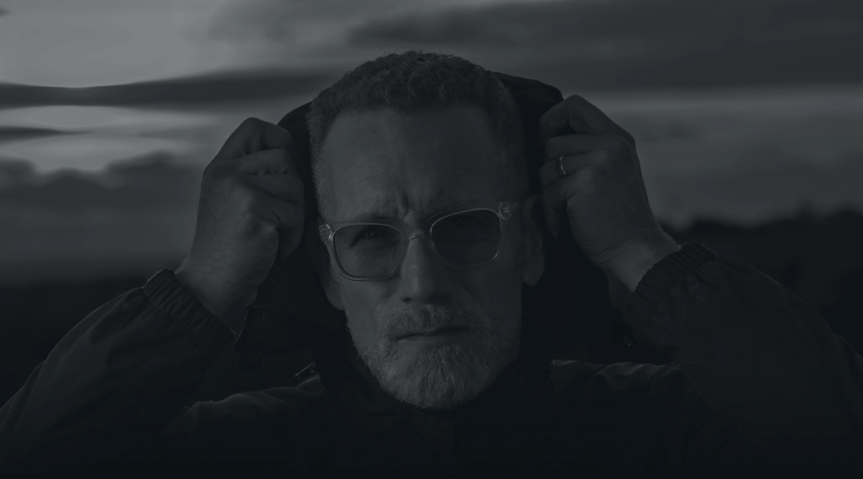 Man with glasses and gray beard pulling up the hood of his jacket outdoors at dusk.