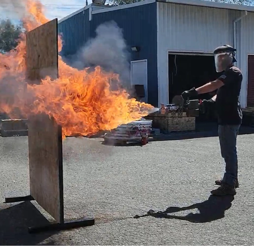 Person wearing protective gear using a flamethrower to project fire at a vertical wooden board outdoors.