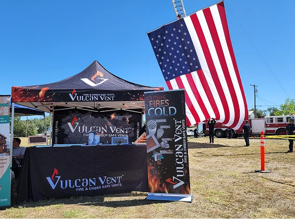 Outdoor booth with Vulcan Vent banners and tent promoting fire and ember safe vents, with a large American flag hanging from a fire truck ladder in the background.