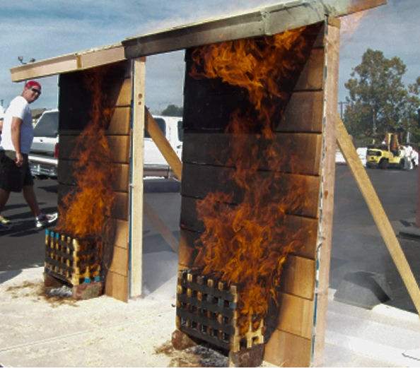 Two vertical wooden structures burning with large flames, with a person walking beside them in an outdoor parking area.