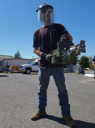 Man wearing a protective face shield and gloves operating a green mechanical device outdoors on a sunny day.