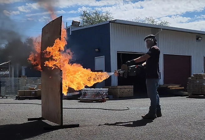 Person using a flamethrower to spray fire at a vertical board outside near a warehouse building.