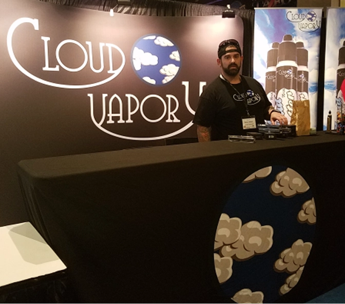 Man standing behind a black booth with Cloud Vapor branding and cloud graphics at a trade show or event.