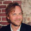 Man with medium-length light brown hair and beard smiling, wearing a dark jacket with a white shirt, standing against a brick and plaster wall.