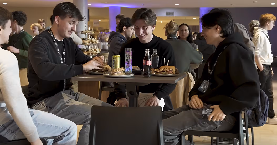 Four young men sitting at a table in a café or dining area, enjoying burgers and drinks while smiling and chatting.