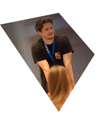 Man wearing a black t-shirt and blue lanyard leaning on a surface, looking at a person with long blonde hair.