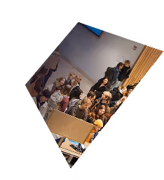 Crowd of people seated and standing indoors, facing a speaker or event out of view.