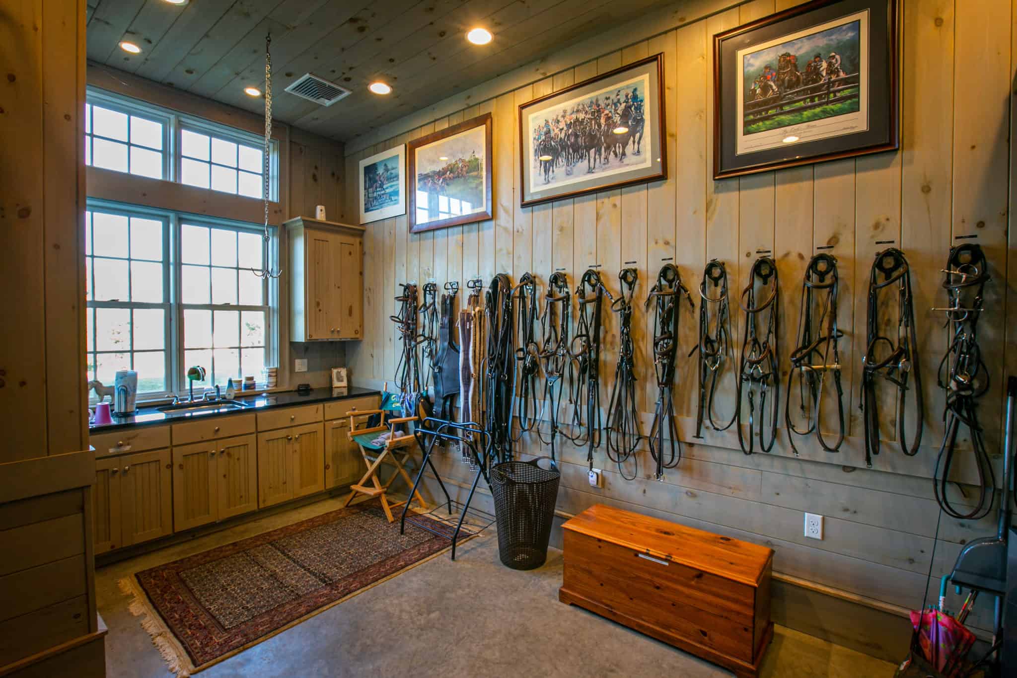 Horse Barn Tack Room with custom cabinetry, rows of bridles hung on a wood-paneled wall and equestrian artwork above.