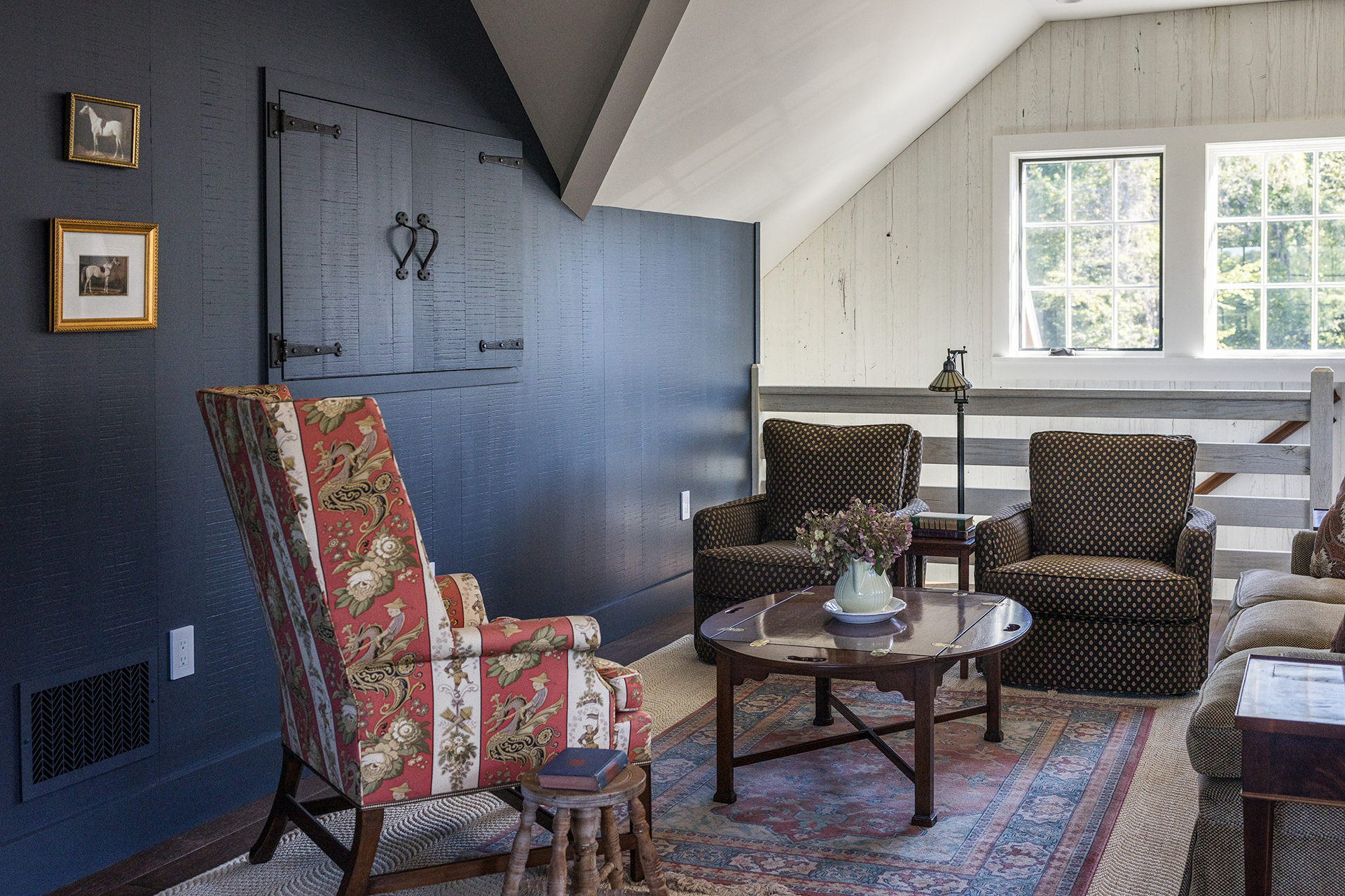 Cozy barn living room with a floral armchair, patterned seating, reclaimed wood shiplap, and equestrian art on navy walls.