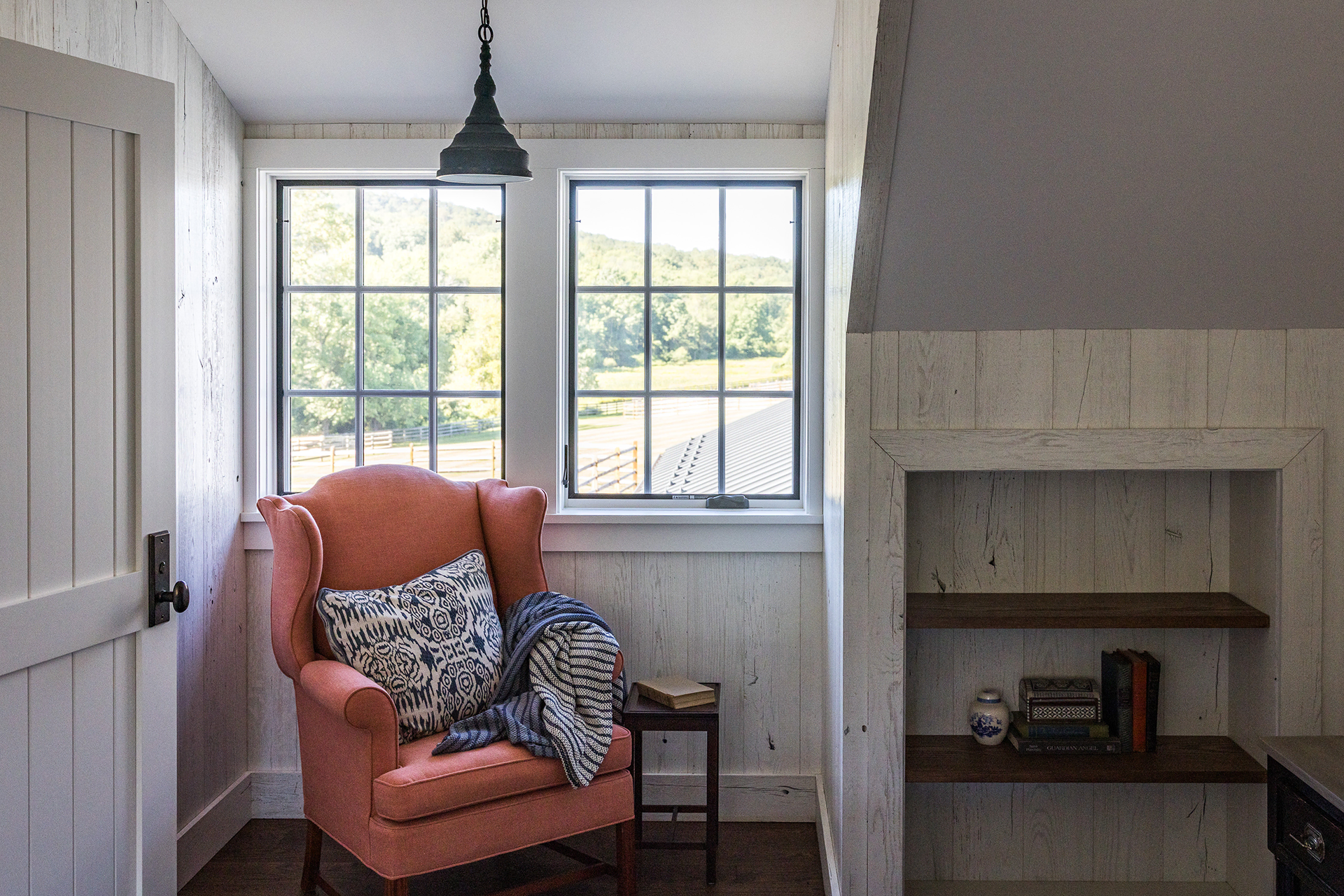 Cozy horse barn living room nook with a coral wingback chair, reclaimed wood shiplap walls, and views of rolling farmland.