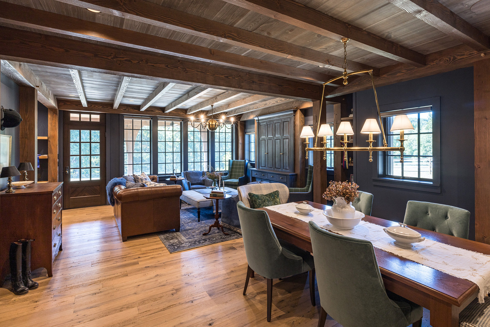 Open-concept lounge and dining space beneath exposed timber frame beams, with large windows and warm wood floors.