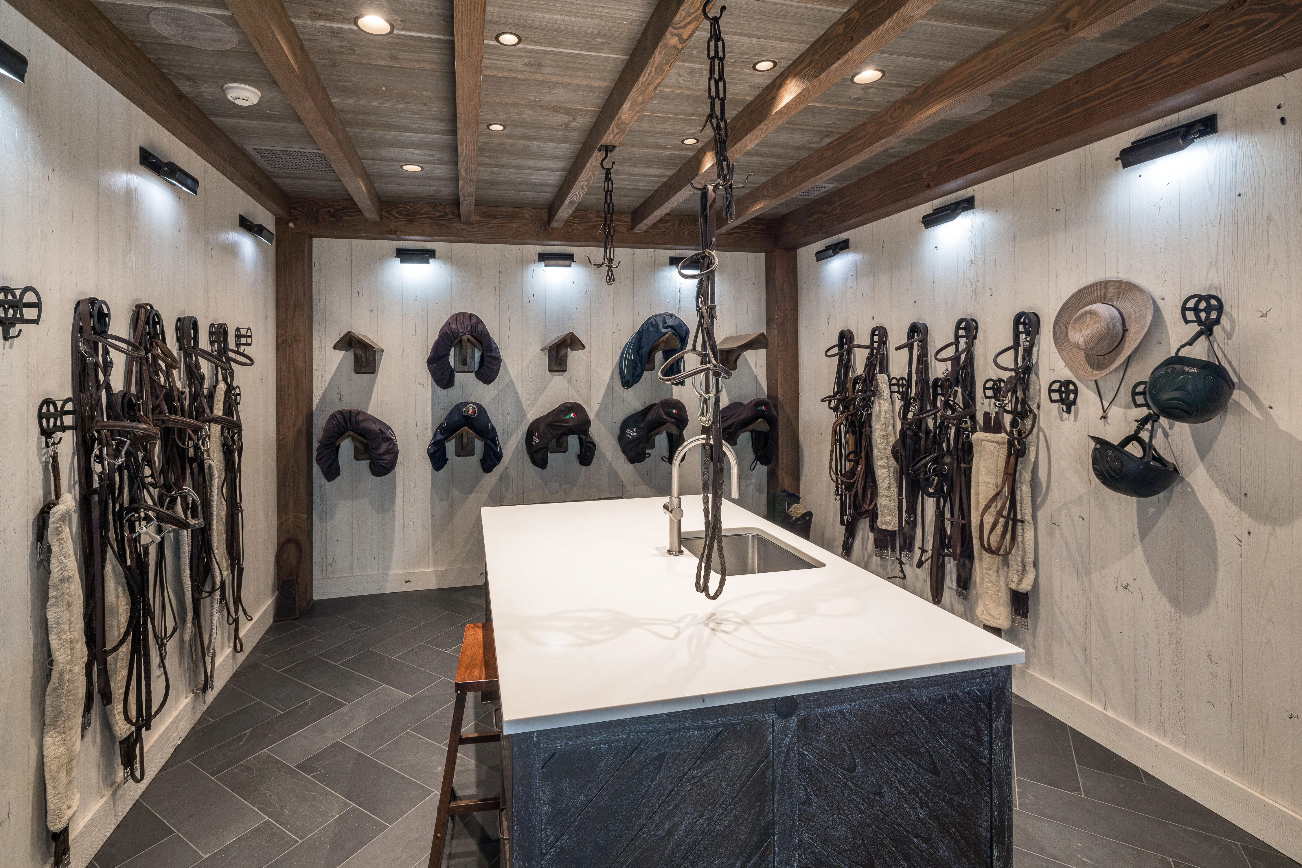 Spacious tack room featuring organization racks for saddles, helmets, and bridles, plus a white granite work island.
