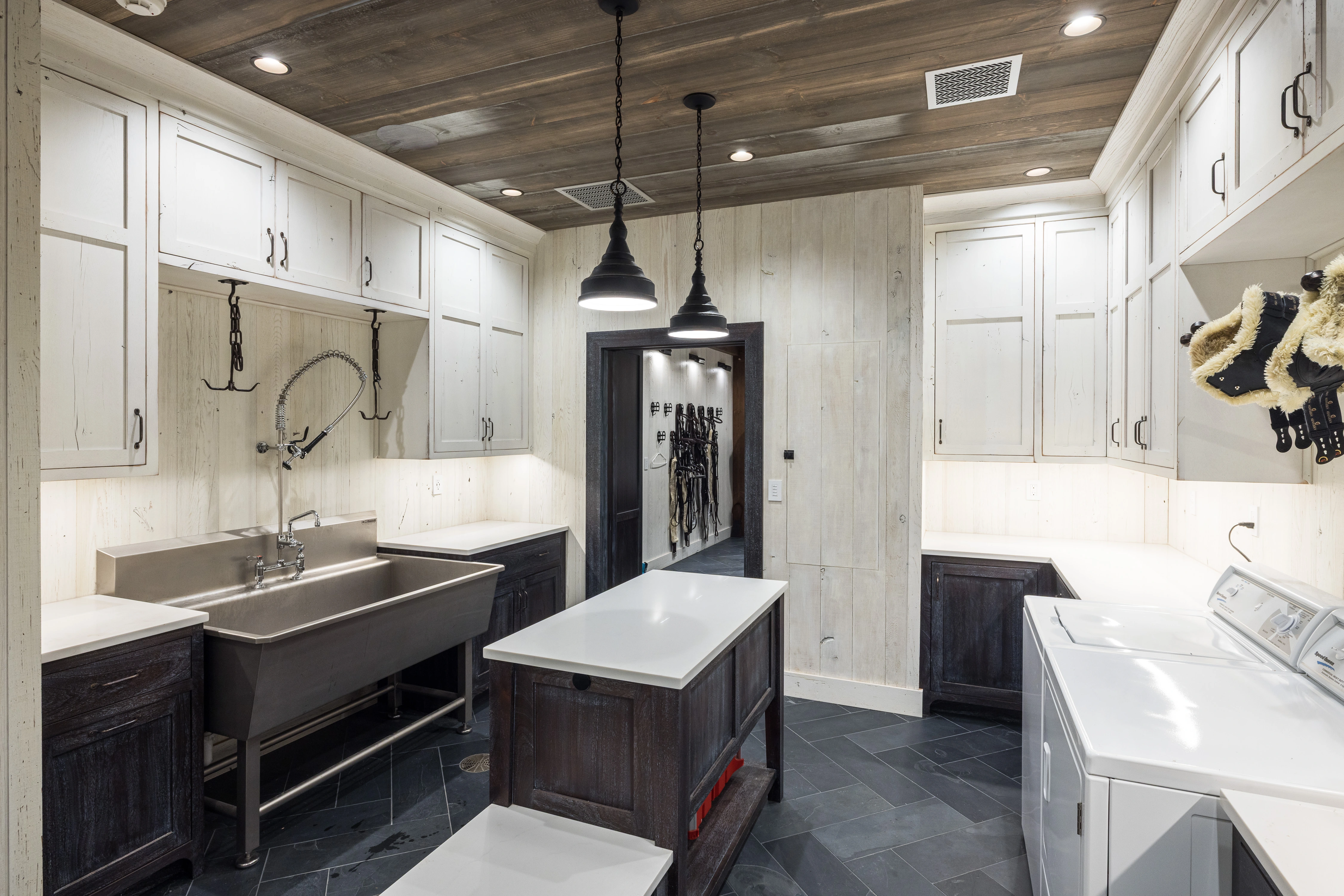 Functional laundry room with custom cabinets, a center island, pendant lights, and an open doorway into the barn’s tack room.