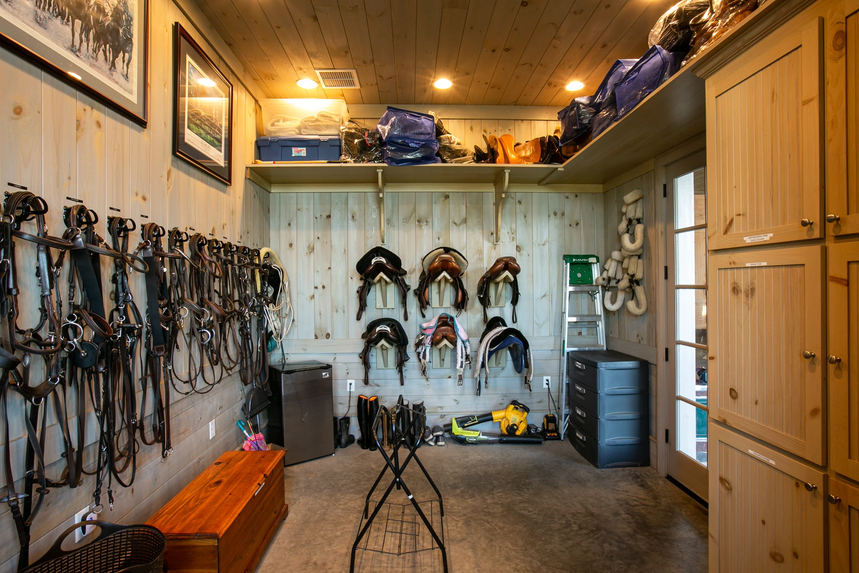 Horse Barn Tack Room with saddles on wall mounted saddle racks, bridles hung on the wall, and built-in storage cabinets.