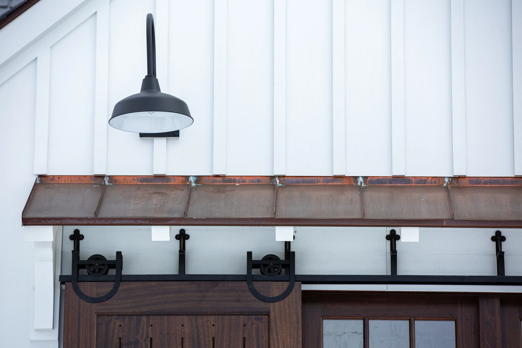 Detail of a copper eave, white board and batten paneling, and black rolling barn door hardware with a black pendant sconce.